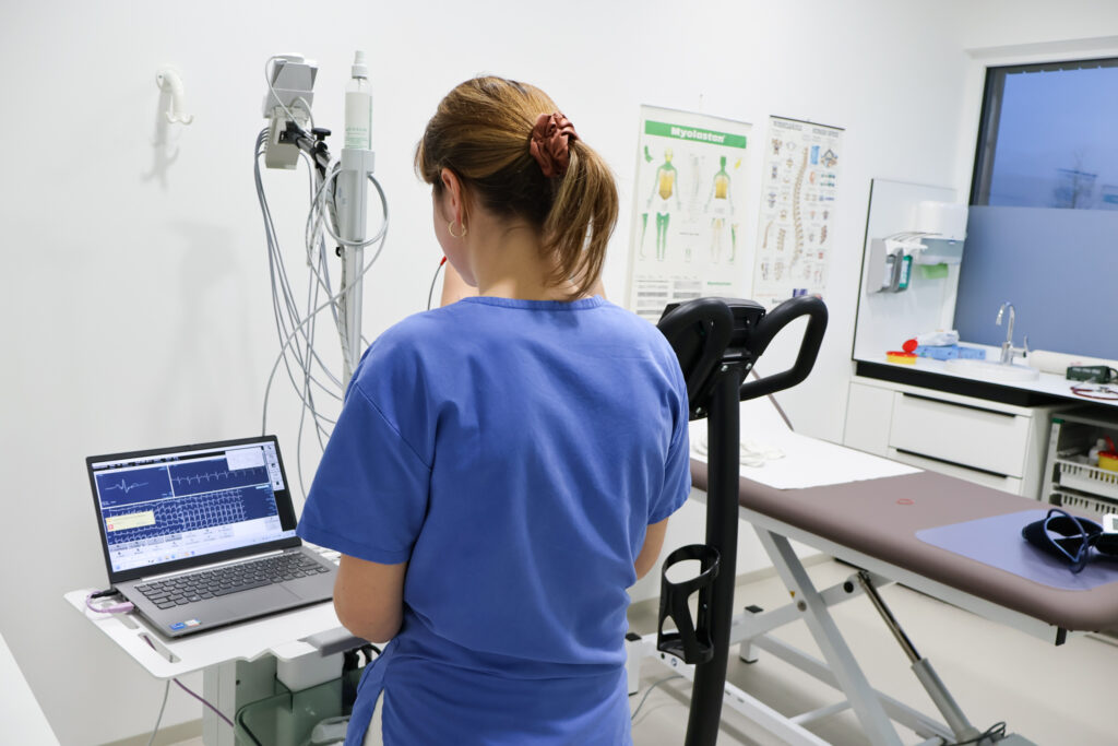 Eine Frau steht vor dem Computer. Sie überwacht die Werte der Person die neben ihr auf dem Rad sitzt und fährt.